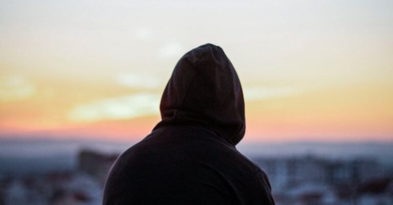 A solitary figure in a hoodie gazes at a vibrant sunset from a rooftop, creating a peaceful silhouette.