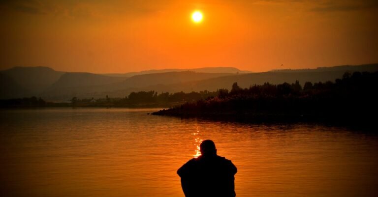 A person sits alone by a tranquil lake, with a stunning sunset in the background.