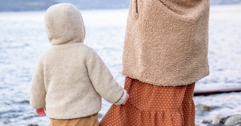 A mother and child in warm clothes walk hand-in-hand by a calm seaside, embracing nature.