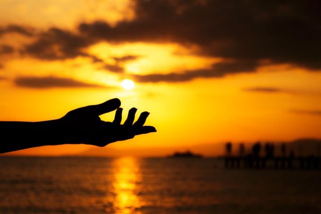A silhouetted hand reaching out towards a vibrant orange sunset over the sea.
